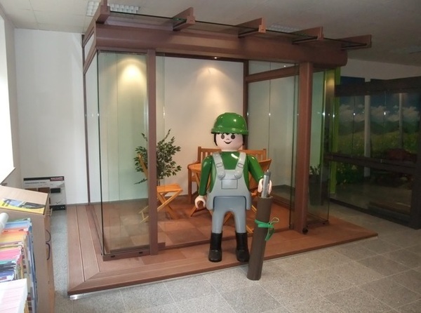 Große Playmobil-Figur steht vor moderner Glasüberdachung mit Gartenmöbeln im Innenraum.